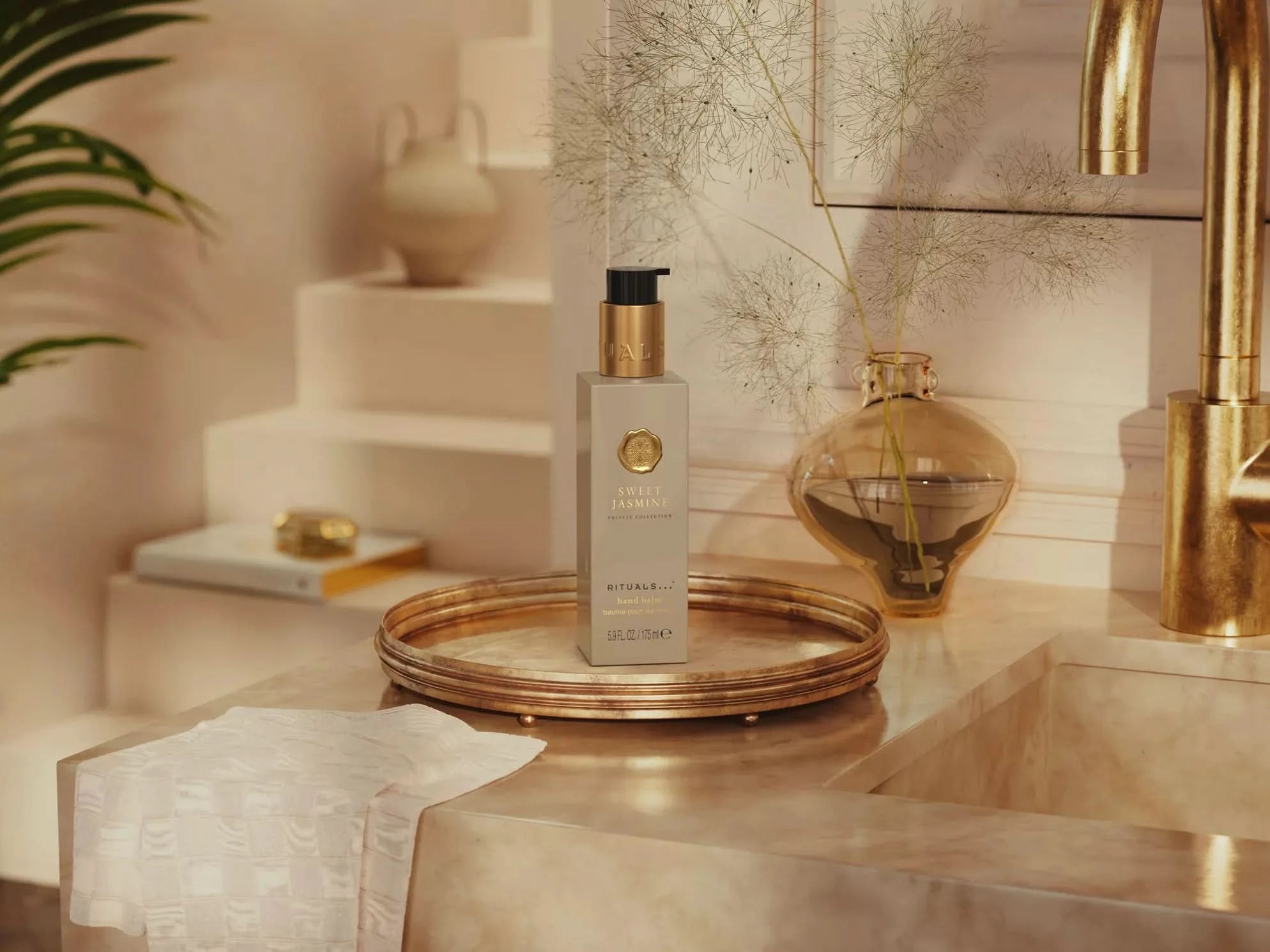 Balsam do rąk Rituals Sweet Jasmine w eleganckiej butelce na miedzianej tacy obok złotego kranu