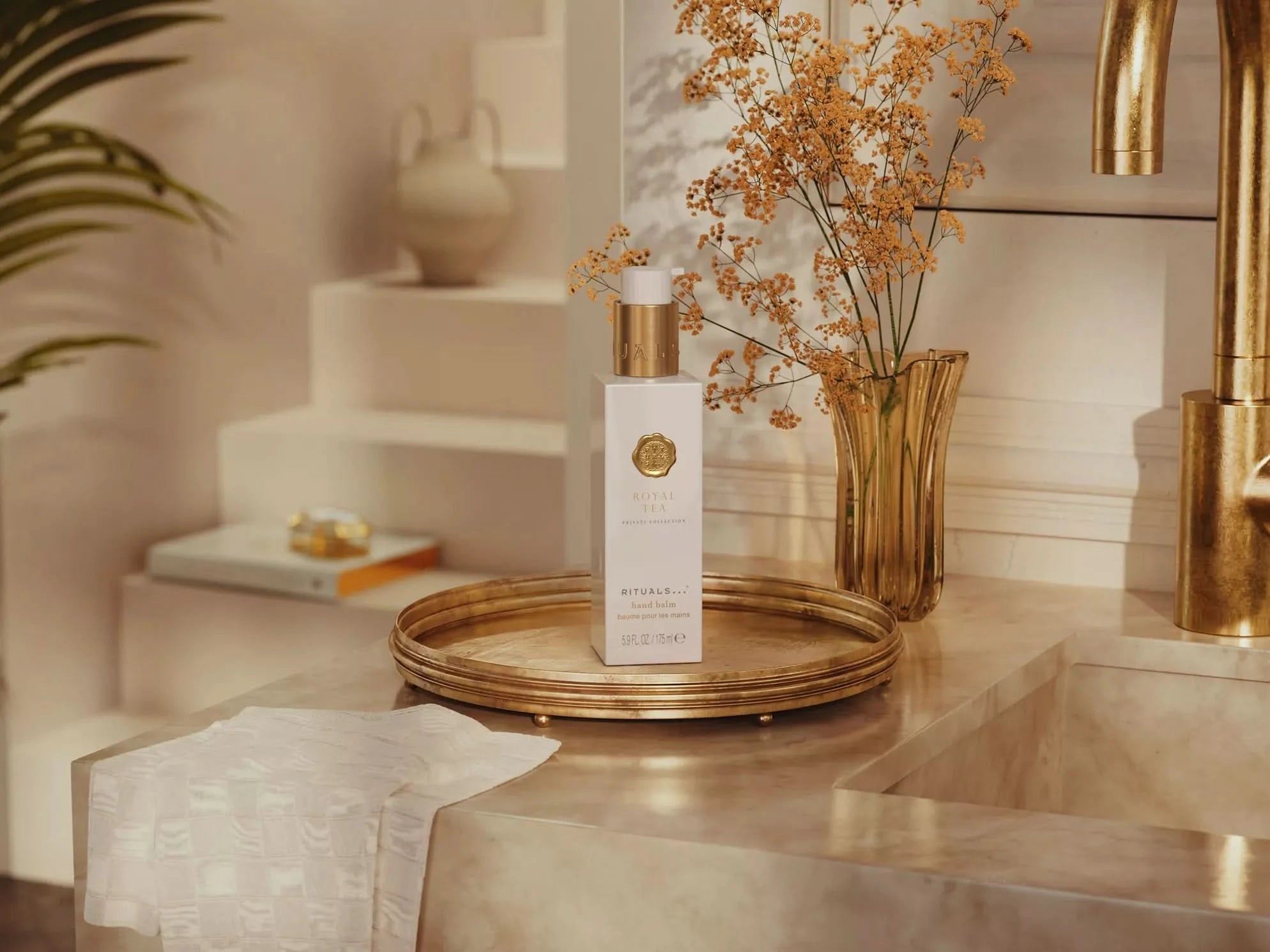 Rituals balsam do rąk Royal Tea na złotej tacy obok wazonu z suszonymi kwiatami przy umywalce