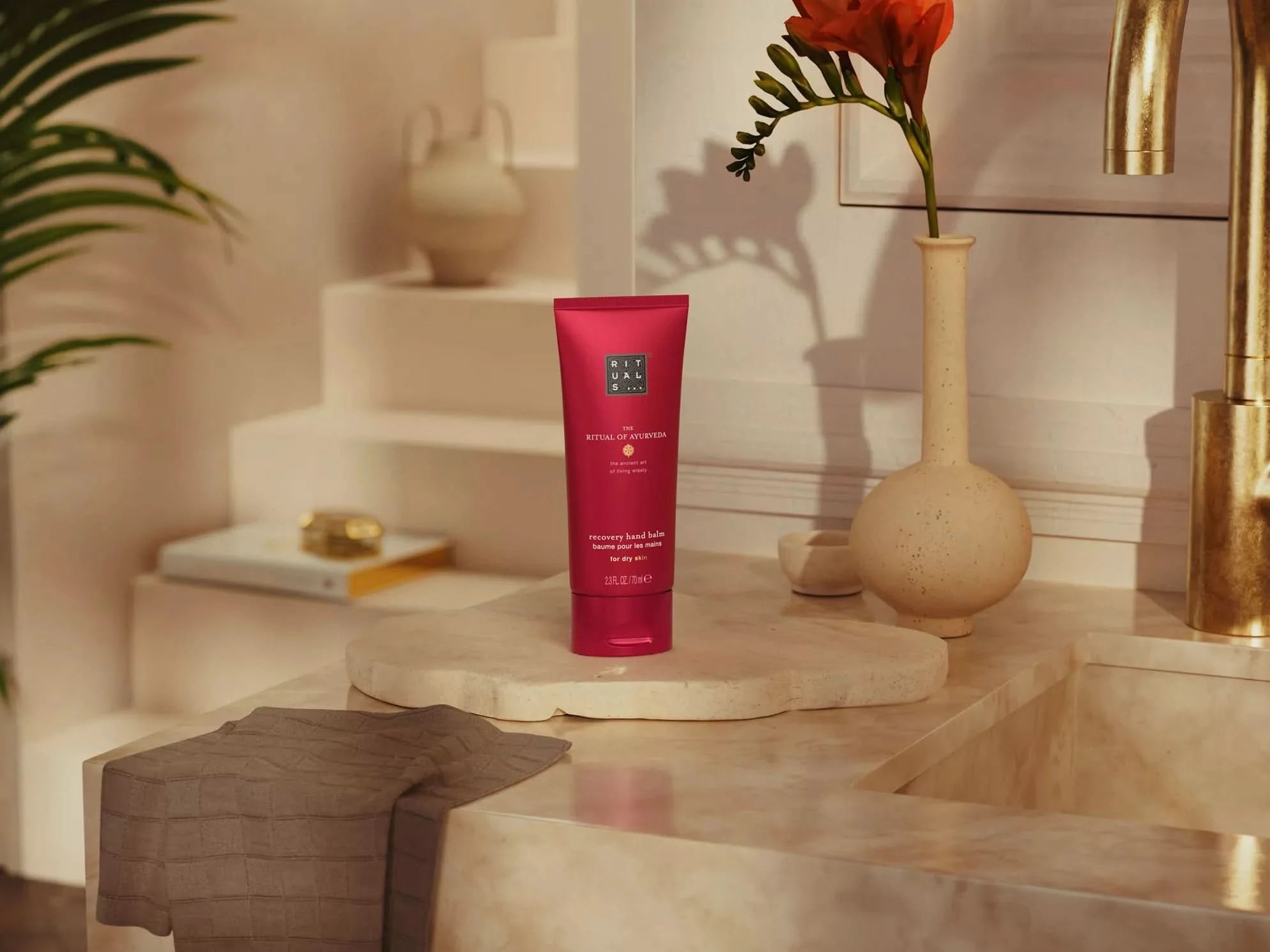 Rituals balsam do rąk w czerwonej tubie na marmurowej łazienkowej półce z kwiatem w wazonie